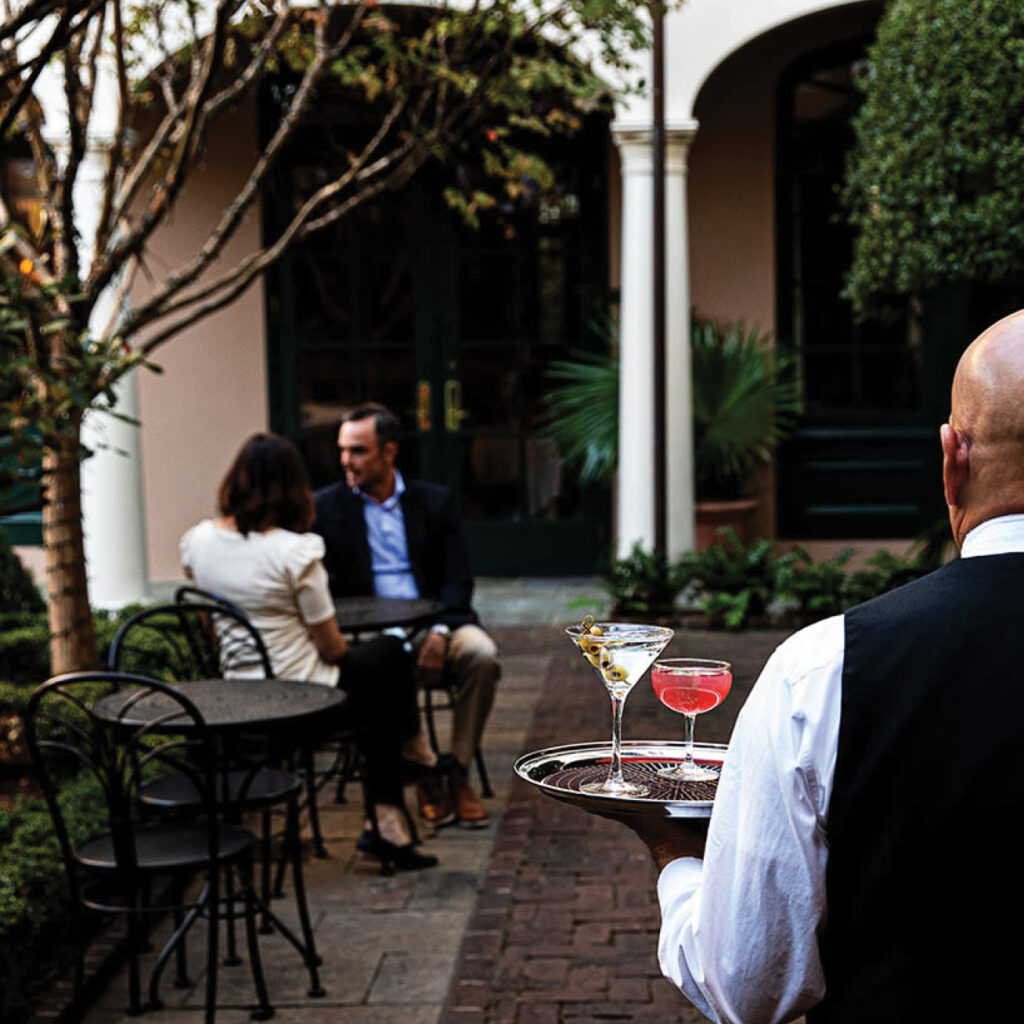 A server holding a tray with two cocktails walking towards a table with two customers in Peninsula Grill's courtyard