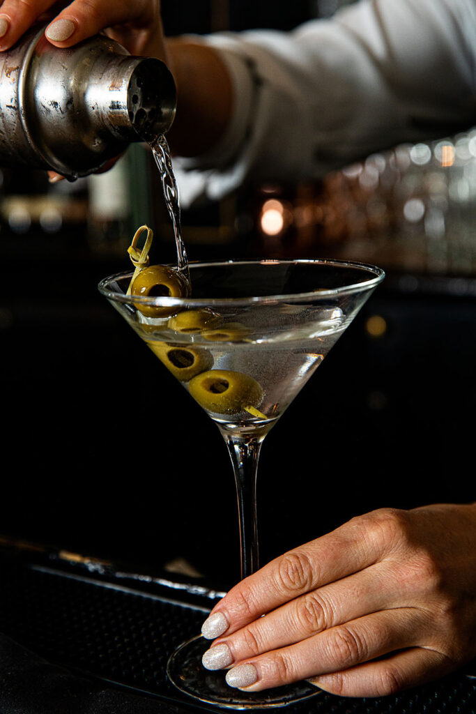 A bartender pouring a martini from a shaker into a martini glass garnished with three green olives