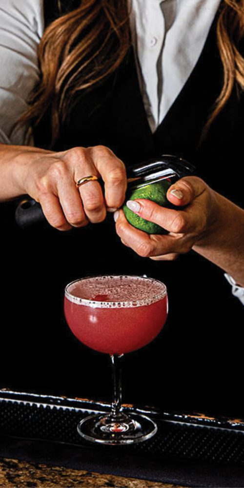 A bartender peeling a lime to garnish a pink cocktail