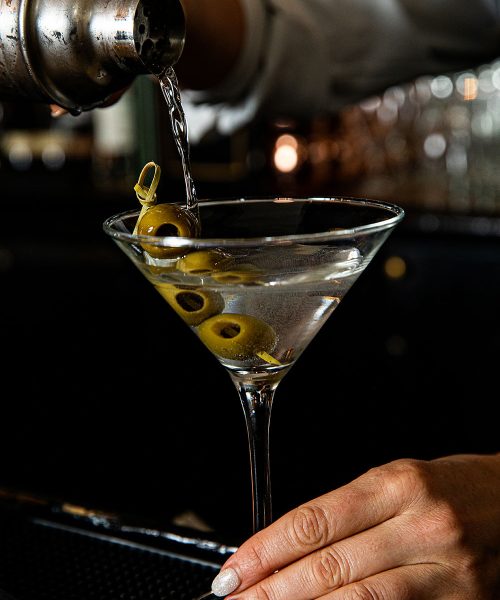 A bartender pouring a martini from a shaker into a martini glass garnished with three green olives